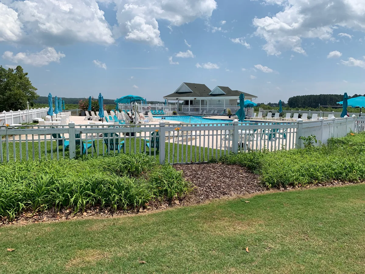 An expansive swimming pool area with lounge chairs, umbrellas, a clubhouse, and a green lawn. Residents are seen relaxing, sunbathing, and socializing, illustrating the leisure amenities available to the community.