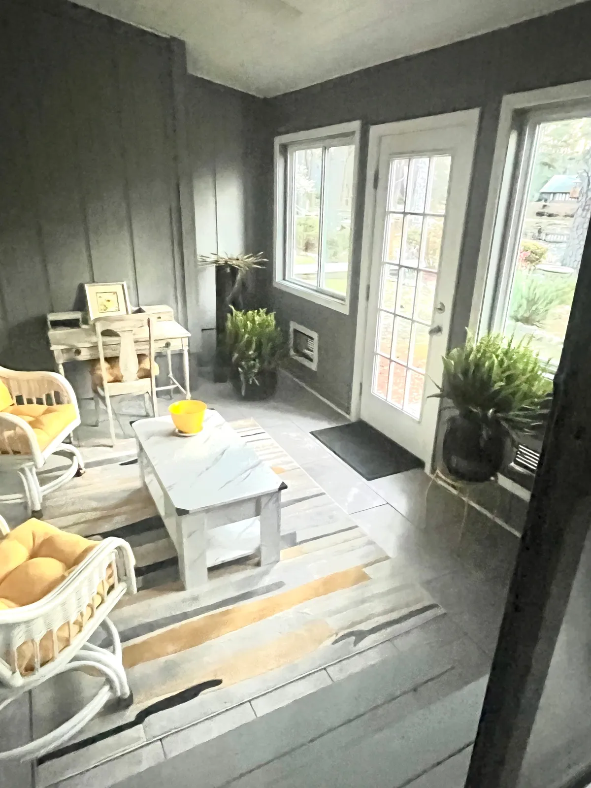 A stylish, comfortable sitting area with wicker chairs, a central coffee table, lush potted plants, and expansive windows that invite in natural light and offer views of the outdoors. The space is perfect for relaxing, reading, or sharing conversations with friends after a day out.