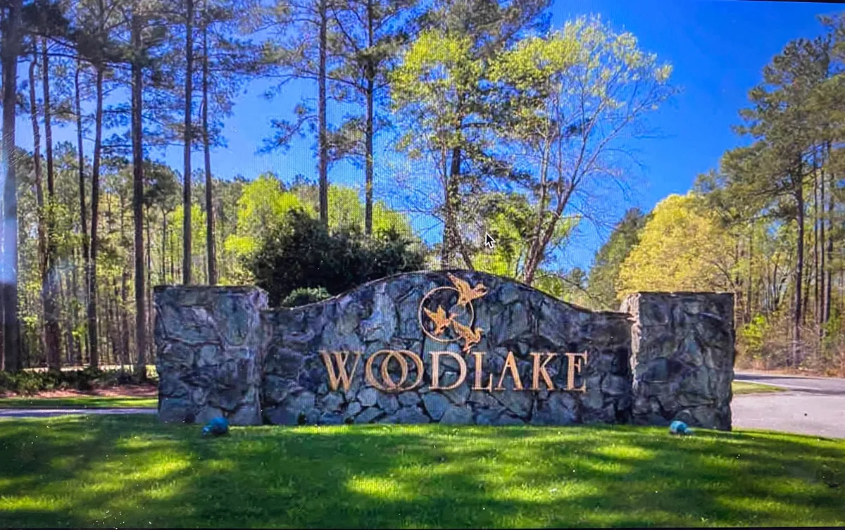 The impressive Woodlake entrance sign framed by landscaped greenery and sturdy stone pillars, with peaceful walking paths inviting residents into a welcoming, secure community.