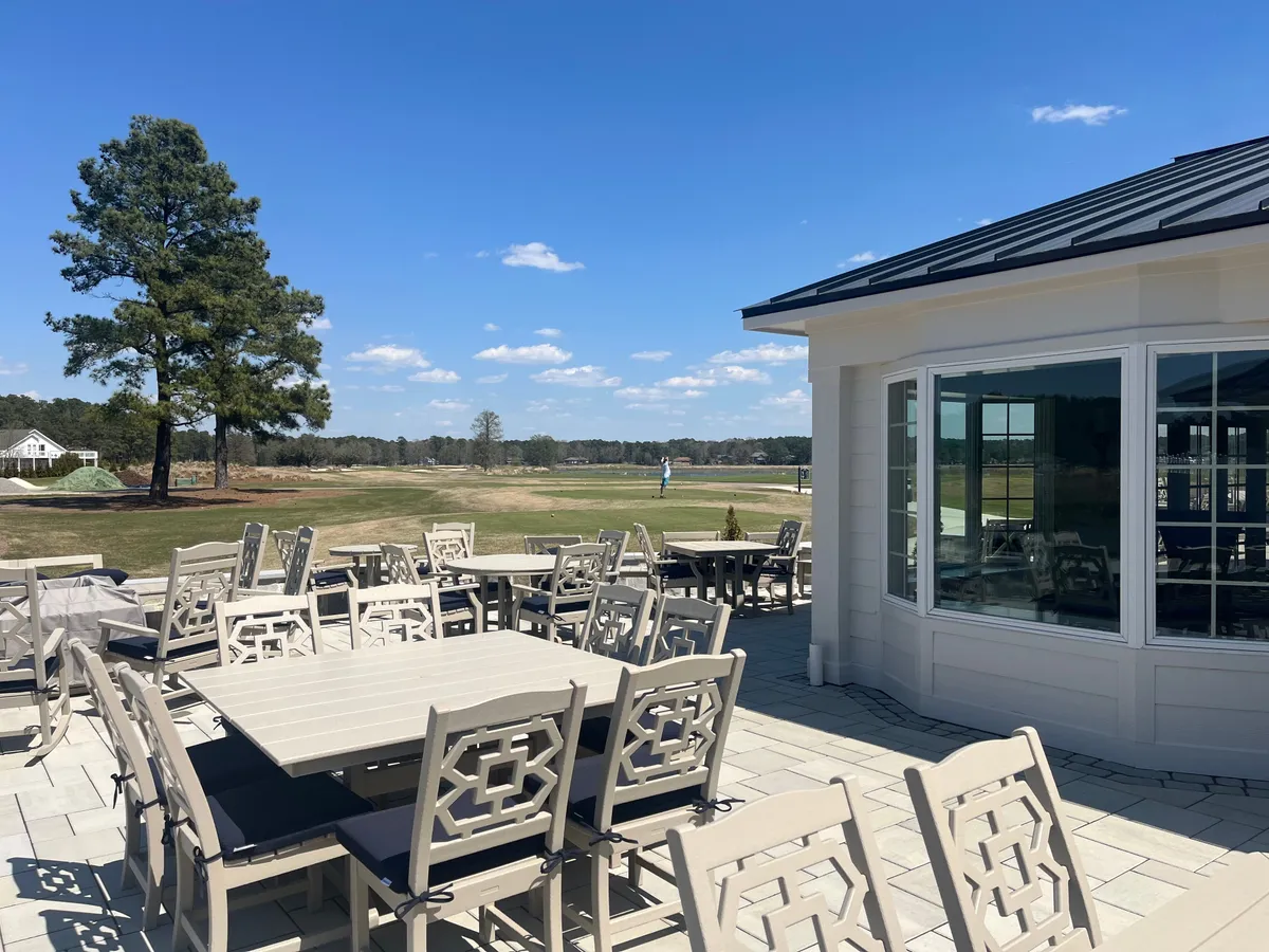 An outdoor seating area overlooks the golf course, with large windows of the building visible behind. The scene exudes a sense of relaxed socializing, al fresco dining, and easy access to the course itself.