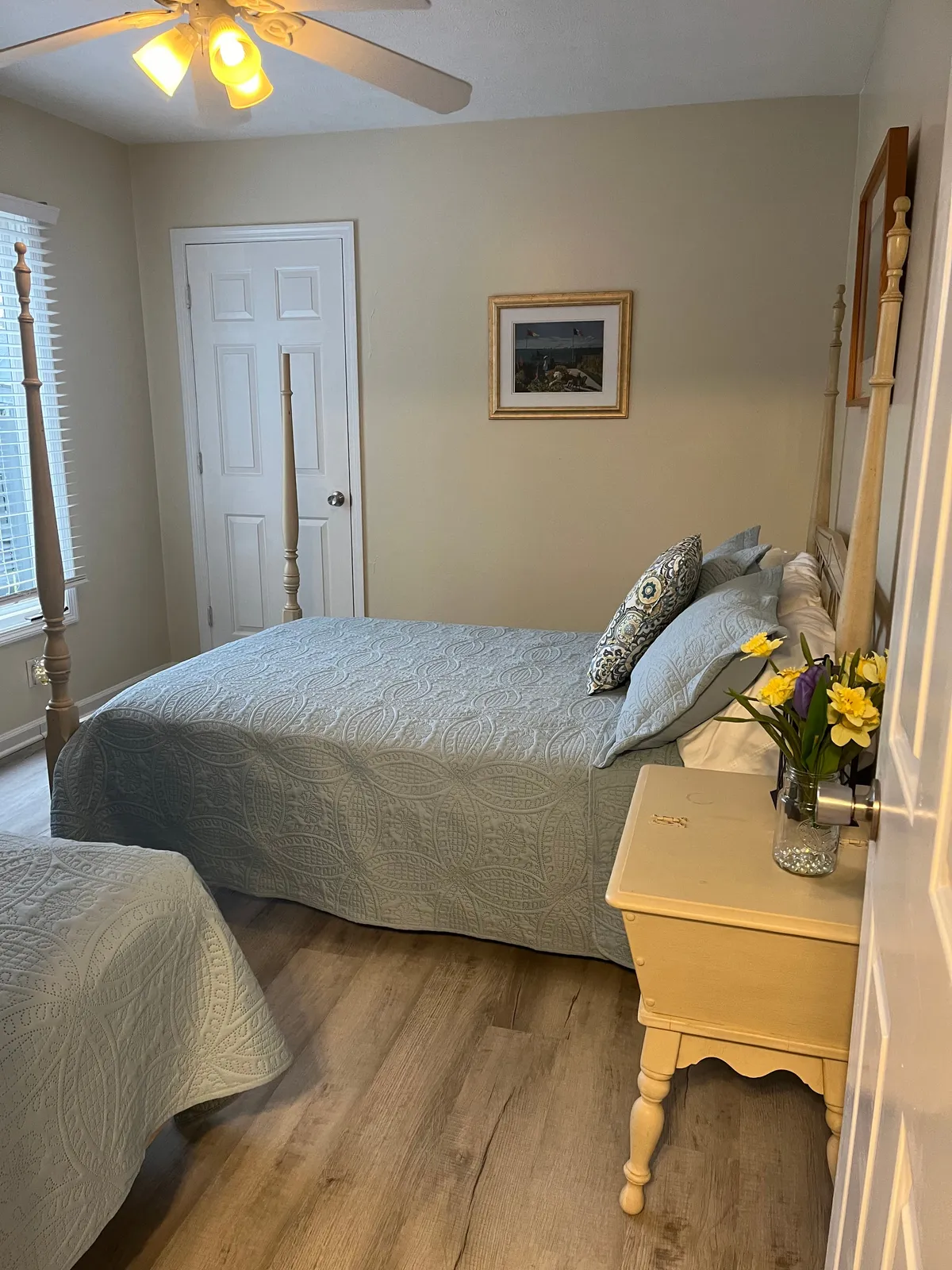A tranquil bedroom scene with a bed adorned in an intricate bedspread, a nightstand topped with fresh flowers, and framed artwork on the wall. Sunlight streams softly into the room, enhancing the serene and restful vibe.