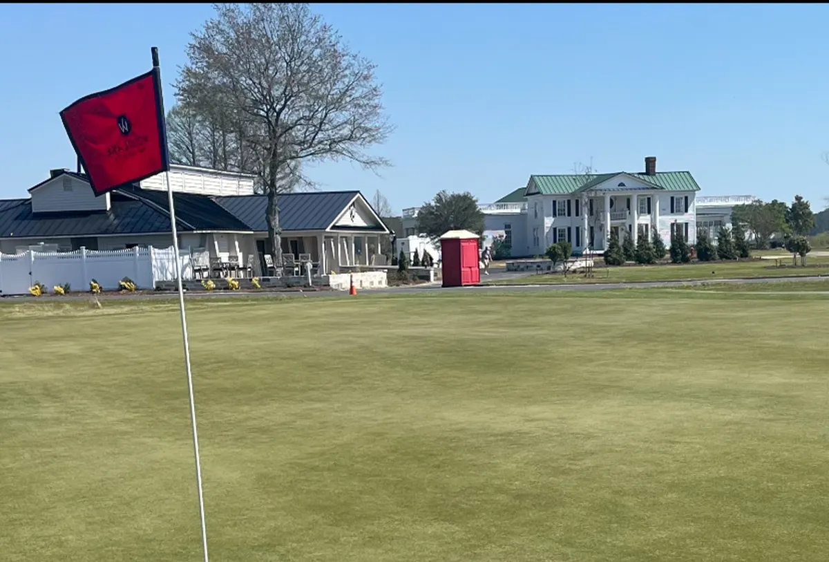 This image features a golf hole with a red flag, a clubhouse, and a large white house in the background. The lush green fairway and relaxed groups of people suggest an exclusive golf community atmosphere, combining both leisure and social opportunities.