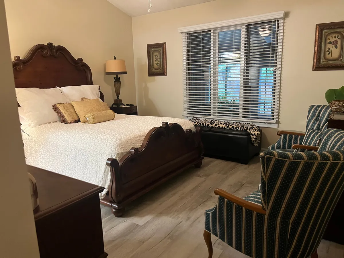 A spacious bedroom featuring an ornate wooden bed with plush bedding, two striped armchairs positioned for conversation or relaxation, a decorative lamp on a nightstand, and framed artwork adorning the walls. Soft, natural light filters in through a nearby window, contributing to the serene ambiance.