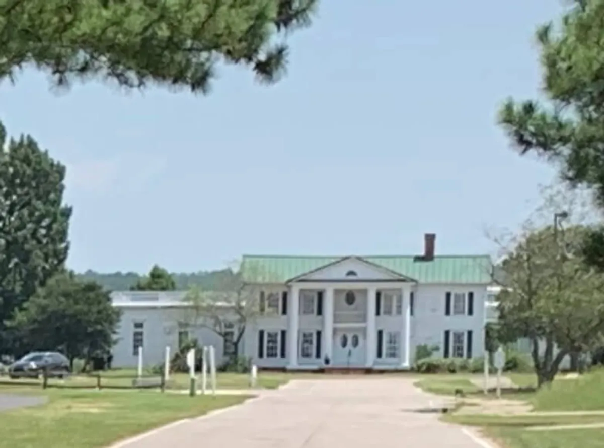 The image captures the grand entrance of the house, featuring a green roof, symmetrical columns, and a welcoming entryway. The scene is framed by lush landscaping, creating an inviting first impression that hints at the security and comfort within the gated community.