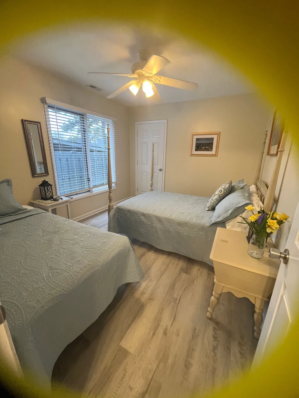 A cozy bedroom featuring two beds with light blue quilts, a shared nightstand with a vase of flowers, and a window with blinds letting in gentle natural light. The room is styled for comfort and shared moments, making it ideal for family or friends.