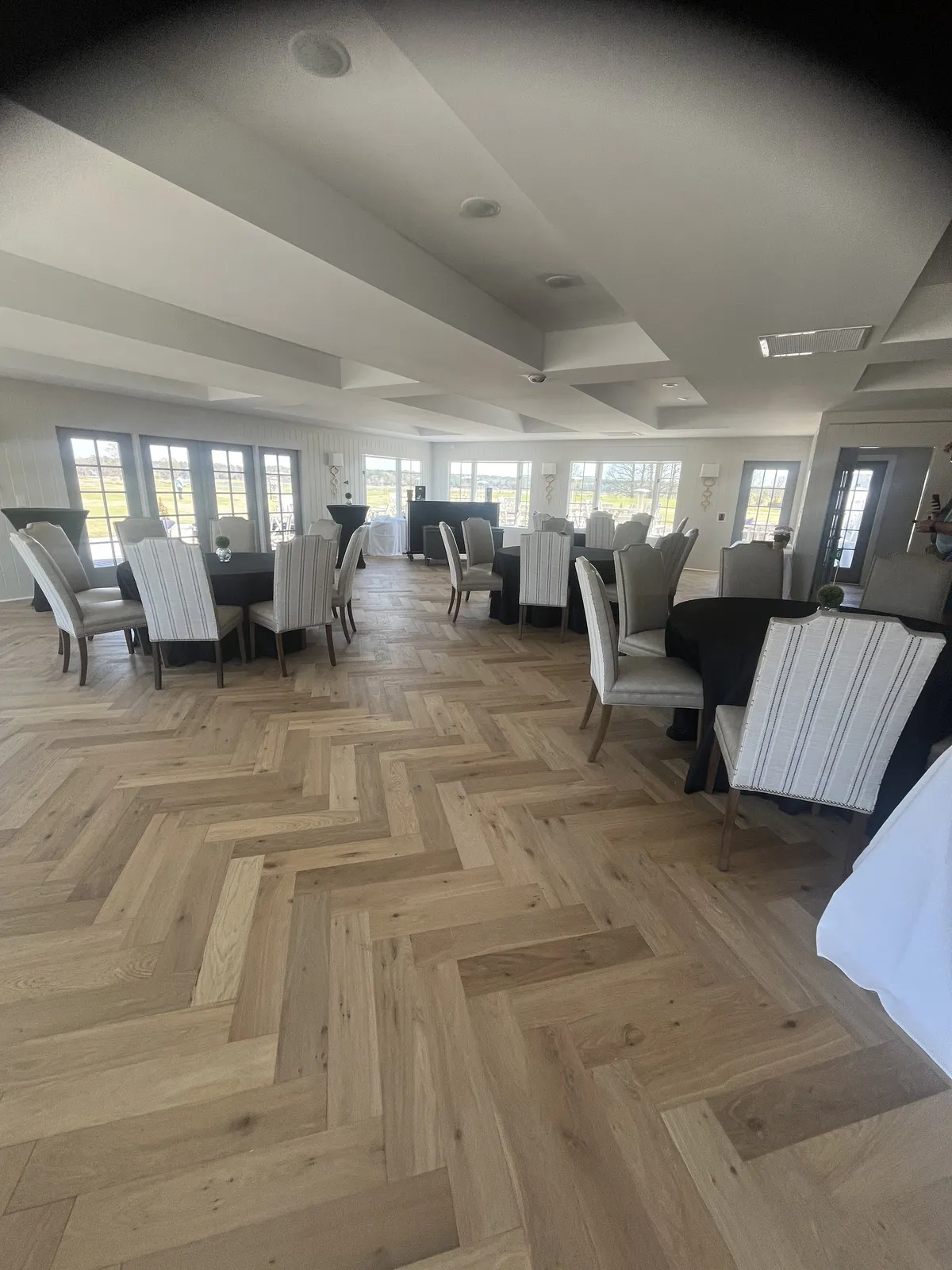 An elegant dining space featuring beautifully set tables, herringbone wood floors, and walls of large windows framing views of the outdoors and flooding the room with natural light. The setting feels sophisticated yet welcoming, ideal for intimate meals and lively social gatherings.