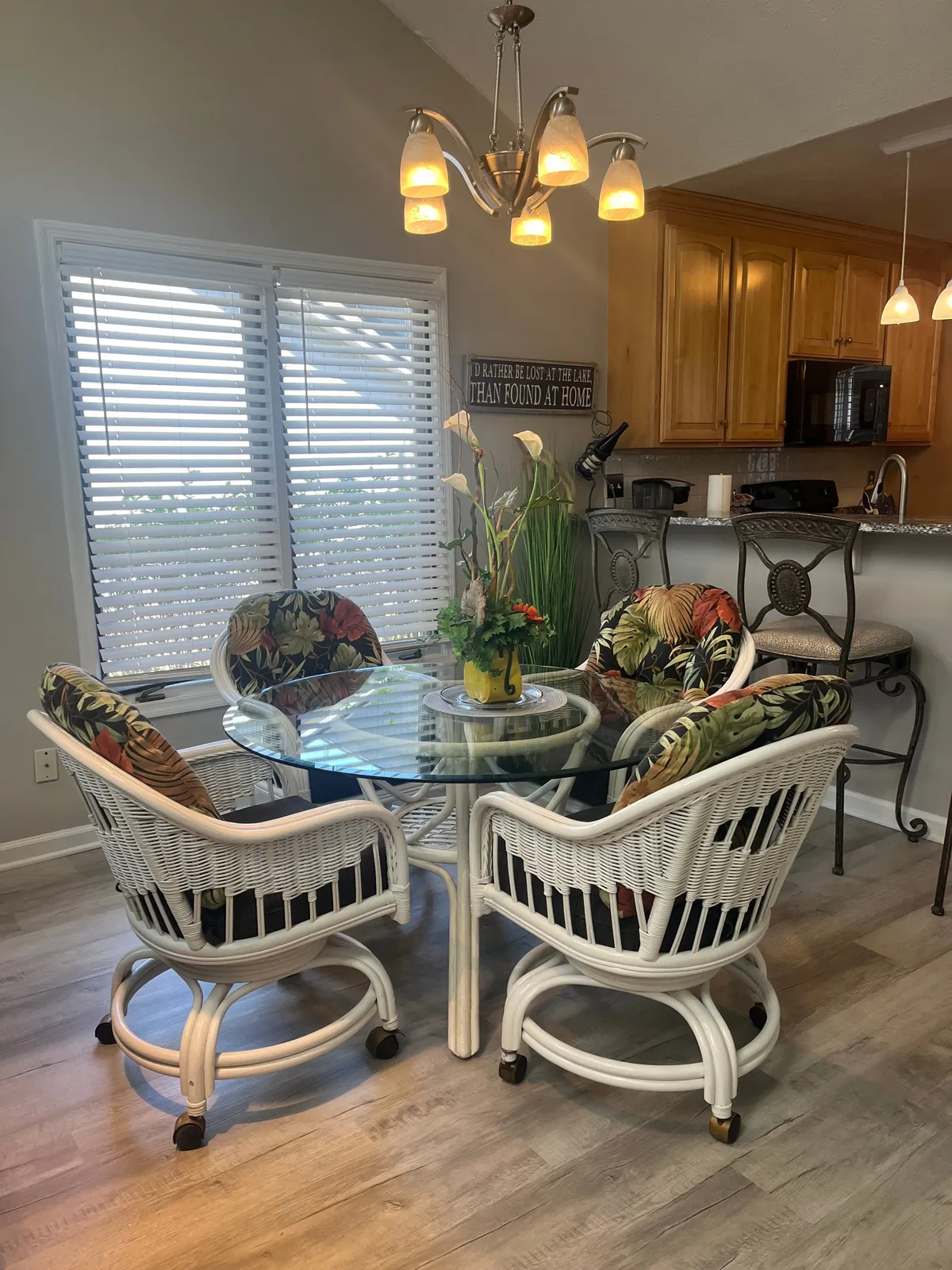A light-filled dining area with a glass table, wicker chairs adorned with floral cushions, and kitchen cabinetry in the background. A decorative plant and stylish light fixture add homely touches, creating a relaxed and welcoming spot for dining or casual conversation.